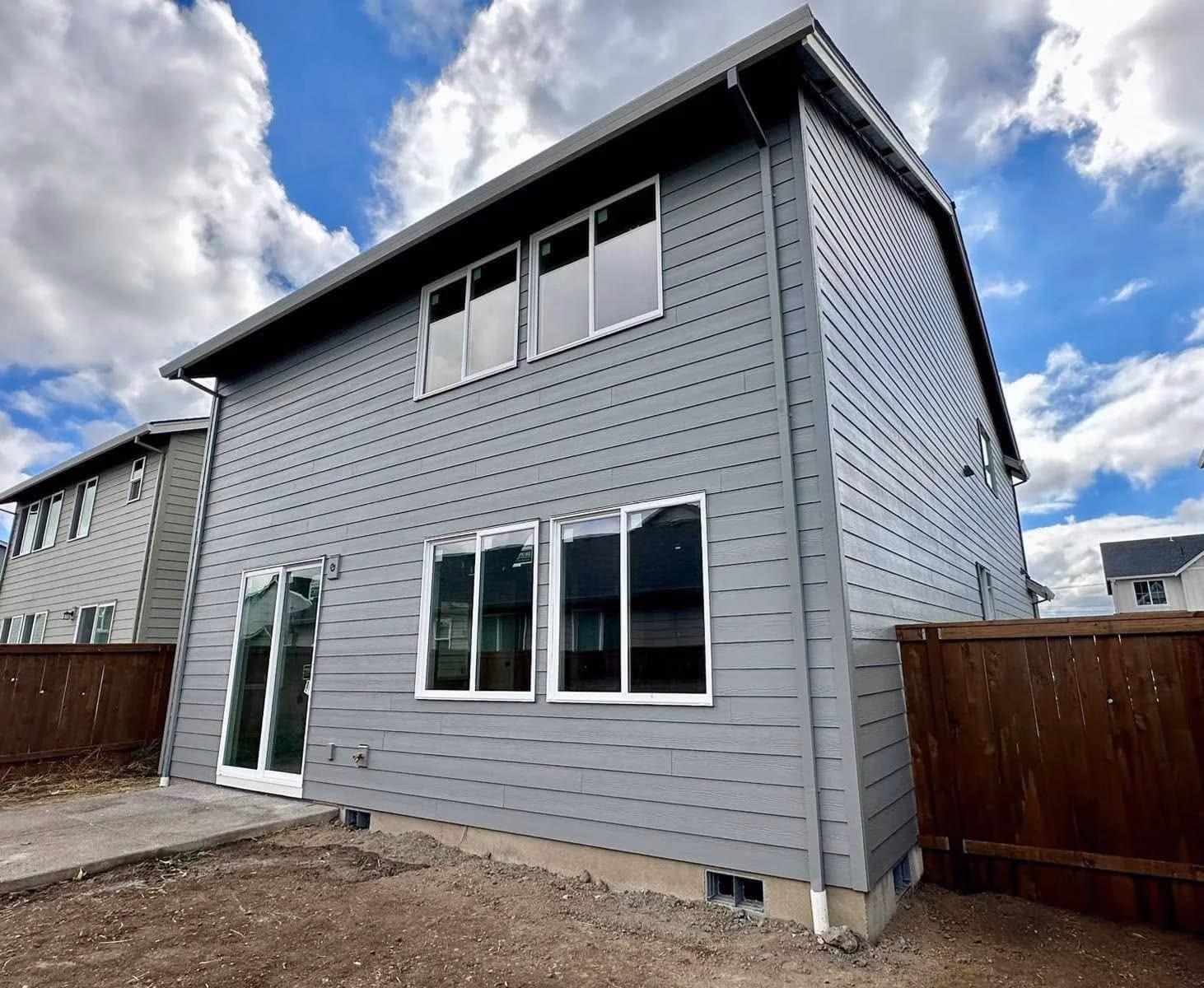 Gray two-story house with white-trim windows and a wooden fence under a partly cloudy sky.