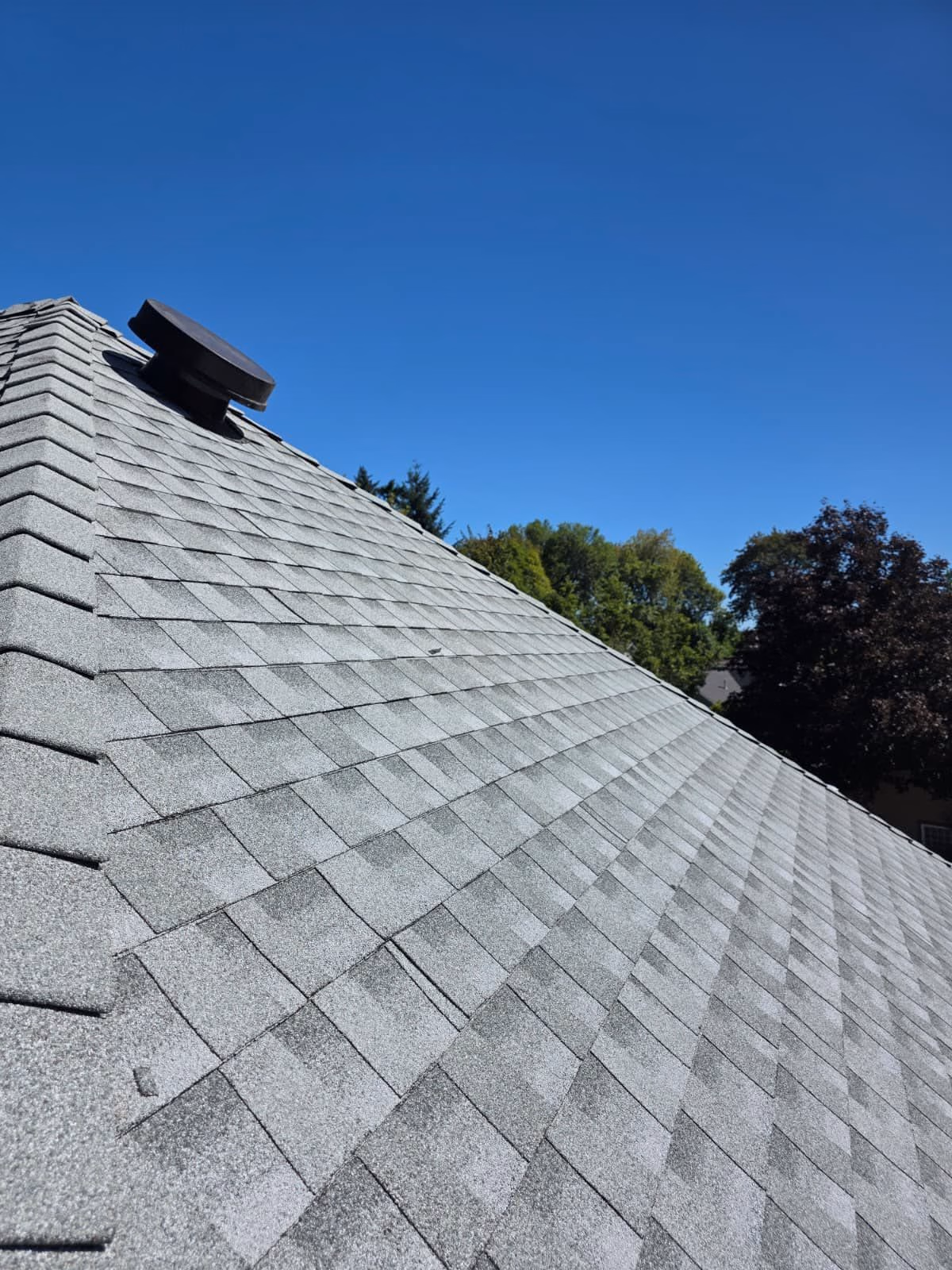 Close-up of a gray shingled rooftop with a black vent pipe and blue sky background.